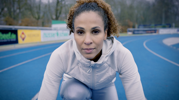 A woman ready to run on a blue running track, looking straight into the camera.